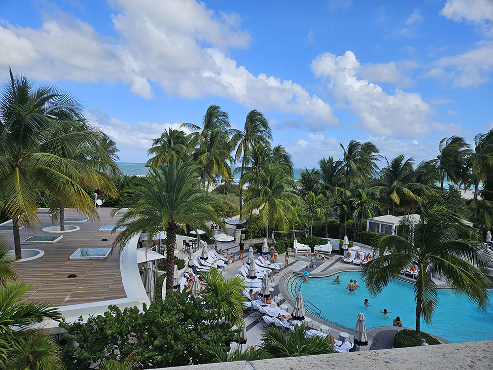 Landscape view of the beach in the distance, with palm trees, the hotel pool and bar in the foreground. Bright blue skies and a few wispy clouds made for an amazing view.